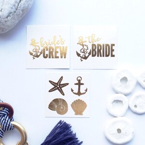 May include: Three sheets of temporary tattoos with a nautical theme. The first sheet says "Bride's Crew" with a gold anchor. The second sheet says "& the Bride" with a gold anchor. The third sheet has a starfish, an anchor, a seashell, and a scallop shell.