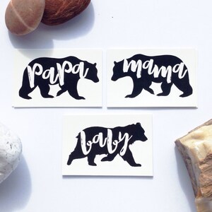 May include: Three black bear silhouette temporary tattoos with the words "papa", "mama", and "baby" written in white inside the bear shapes.