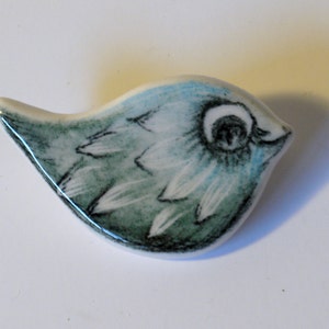 Könnte beinhalten: Eine Keramik-Brosche in Form eines Vogels mit einem blauen und grünen bemalten Design. Der Vogel hat ein einzelnes Auge und ein detailliertes Federmuster.