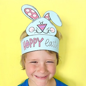 May include: A child wearing a blue shirt and a paper bunny headband that says "Hoppy Easter" with a pink egg graphic. The headband has pink ears with white polka dots.
