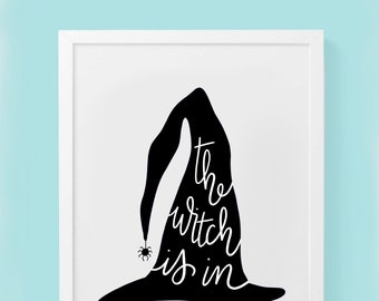 May include: A framed print featuring a black witch's hat silhouette with the phrase "the witch is in" in white script. A small spider graphic is near the hat's brim. The artwork is set against a white background.