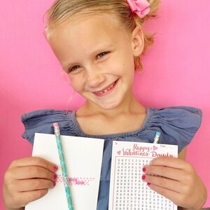 May include: A young girl holds a pink and white Valentine's Day card with a word search puzzle. The card says "Happy Valentines Day" and has a heart with the word "from" and a heart with the word "to". The word search puzzle has the words "amazing, awesome, cool, fabulous, fantastic, magnificent, spectacular, terrific, wonderful".