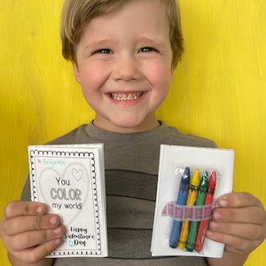 May include: A child holds a Valentine's Day card that says "You color my world!" and a small package of crayons. The card is decorated with a heart and the text "Happy Valentine's Day".