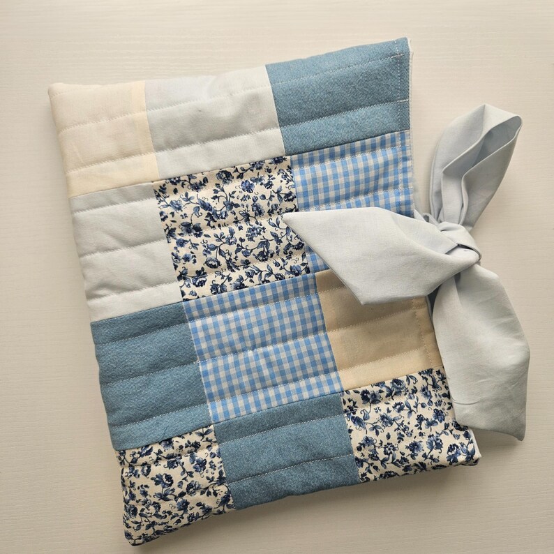 May include: A patchwork cushion with squares of light blue, cream, and floral fabric. The cushion is tied with a light blue bow. The cushion is approximately 30 cm square.