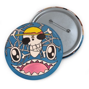 May include: A blue and white button with a cartoon skull wearing a straw hat with a cross on it. The skull is surrounded by a black web-like design. The button has a pink and white mouth with sharp teeth.