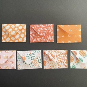 Op de afbeelding: Een verzameling van zeven kleine, decoratieve papieren enveloppen. De enveloppen hebben verschillende patronen, waaronder oranje en witte stippen, bloemmotieven en geometrische vormen. Het kleurenpalet omvat oranje, roze, groen en wit.
