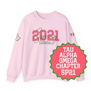 May include: A light pink sweatshirt with the text "Spring 2021" in pink and green script. The sweatshirt also features a green and pink emblem on the sleeve and a red and green badge that says "Tau Alpha Omega Chapter SP21".