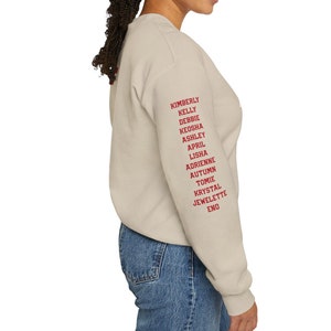 May include: A light beige sweatshirt with long sleeves and a crew neck. The right sleeve has a list of names printed in red: Kimberly, Kelly, Debbie, Keosha, Ashley, April, Lisha, Adrienne, Autumn, Tomie, Krystal, Jewlette, Eno.