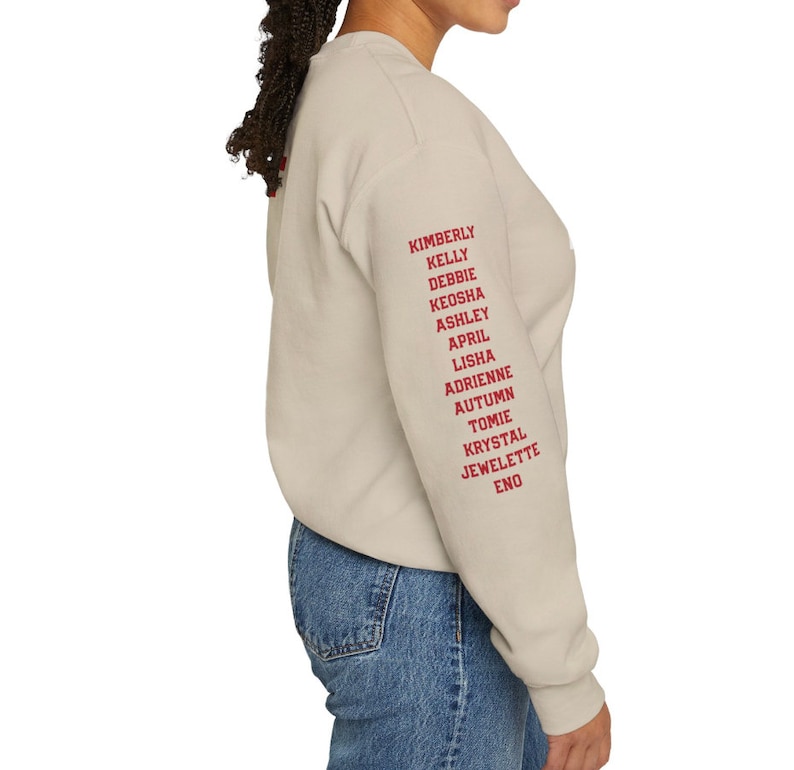 May include: A light beige sweatshirt with long sleeves and a crew neck. The right sleeve has a list of names printed in red: Kimberly, Kelly, Debbie, Keosha, Ashley, April, Lisha, Adrienne, Autumn, Tomie, Krystal, Jewlette, Eno.