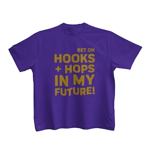 May include: A purple t-shirt with the text "BET ON HOOKS + HOPS IN MY FUTURE!" in gold lettering. The shirt is a crew neck style and appears to be made of cotton or a similar fabric. The text is centered on the front.