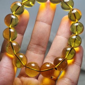 May include: A beaded bracelet featuring large, translucent amber-colored beads. The beads are round and strung together, creating a warm, golden glow. The bracelet is held in a hand, showcasing the natural color and clarity of the stones.