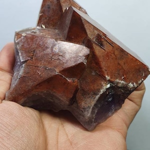 May include: A large, faceted amethyst crystal with a deep purple hue and a reddish-brown tint. The crystal has a rough, natural texture and is shaped like a pyramid.