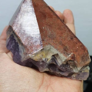 May include: A large, amethyst crystal with a reddish-brown, layered exterior. The crystal has a pointed top and a rough, uneven base.