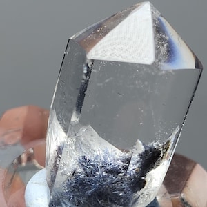 May include: A clear quartz crystal with a pointed tip and dark blue needle-like inclusions. The crystal sits on a white base, highlighting its transparency and internal features. The background is a soft grey.