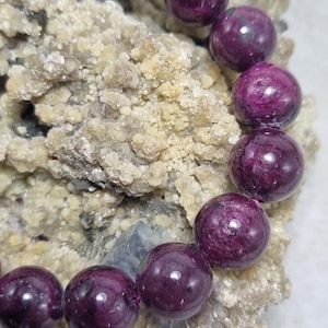 May include: A bracelet featuring large, round, deep purple beads. The beads are a rich, dark color with a glossy finish. The bracelet is resting on a textured, light-colored surface, creating a contrast with the beads.
