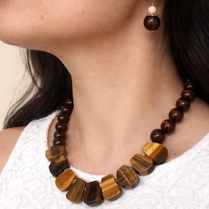 以下が含まれることがあります： 濃い茶色の木製ビーズとタイガーアイストーンのペンダントが特徴のネックレスとイヤリングのセット。ネックレスは丸いビーズと平らな幾何学的なペンダントが連なっています。イヤリングも同様のデザインです。