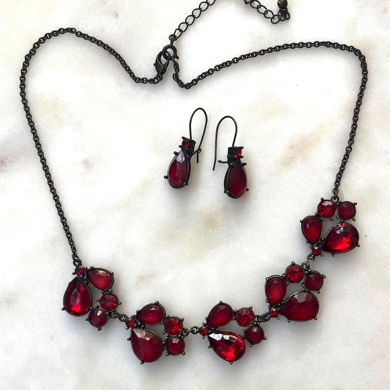Pu&ograve; includere: Un set di collana e orecchini in metallo canna di fucile scuro. La collana presenta una serie di pietre rosse a forma di goccia. Gli orecchini abbinati hanno un design simile. I gioielli sono su uno sfondo di marmo bianco.