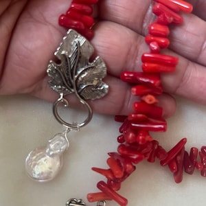 Puede incluir: Un collar de coral rojo brillante con un colgante de hoja plateada y una perla grande e irregular. El collar est&aacute; compuesto por peque&ntilde;as piezas de coral rojo vibrante. El colgante es una hoja plateada detallada con una gran gota de perla.