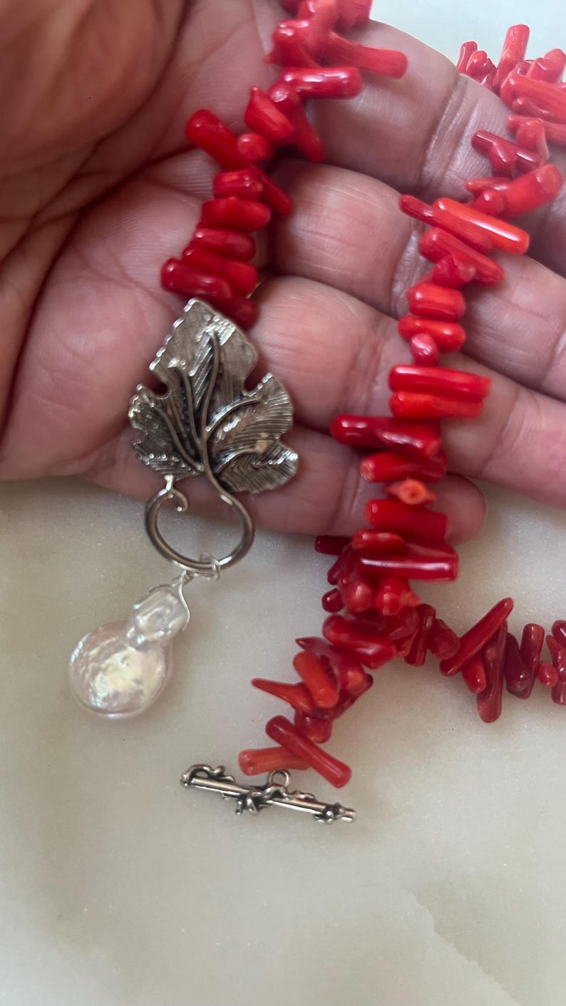 Puede incluir: Un collar de coral rojo brillante con un colgante de hoja plateada y una perla grande e irregular. El collar est&aacute; compuesto por peque&ntilde;as piezas de coral rojo vibrante. El colgante es una hoja plateada detallada con una gran gota de perla.