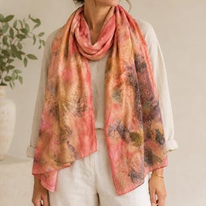 May include: A rectangular scarf featuring a botanical print with shades of coral, pink, and muted earth tones. The fabric displays detailed leaf imprints in dark brown and green scattered across the surface, draped loosely around the neck.