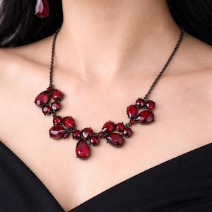 Pu&ograve; includere: Una collana e orecchini abbinati rosso scuro. La collana presenta pietre preziose a forma di goccia e ovali rosso scuro incastonate in una catena di metallo scuro. Gli orecchini hanno un design simile, con una singola pietra preziosa a goccia.