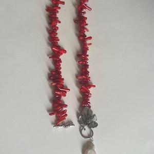 Puede incluir: Un collar de coral rojo brillante con un cierre plateado y un colgante en forma de hoja. El colgante sostiene una gran perla en forma de l&aacute;grima. El collar est&aacute; hecho de peque&ntilde;os trozos irregulares de coral.
