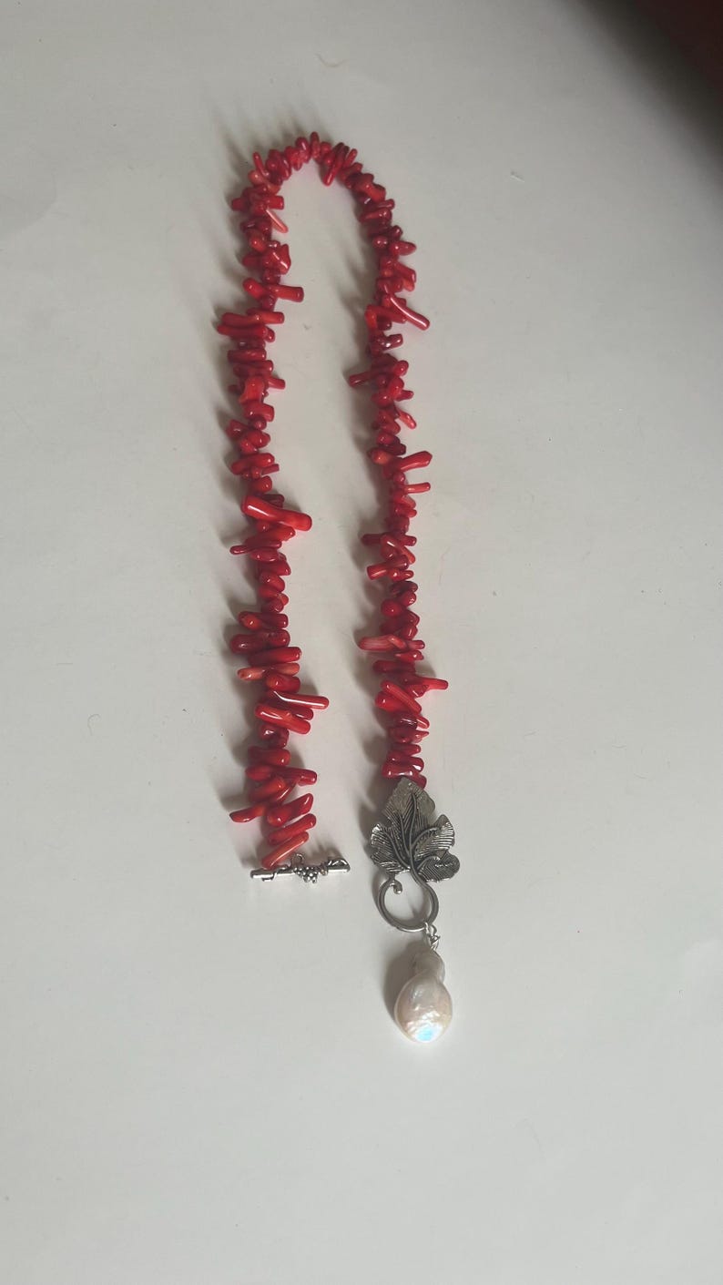 Puede incluir: Un collar de coral rojo brillante con un cierre plateado y un colgante en forma de hoja. El colgante sostiene una gran perla en forma de l&aacute;grima. El collar est&aacute; hecho de peque&ntilde;os trozos irregulares de coral.