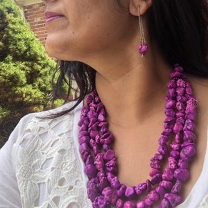 Pode incluir: Um colar de várias camadas e brincos a condizer com pedras roxas irregulares e vibrantes. O colar tem várias camadas de pedras, criando um acessório arrojado e apelativo. Os brincos penduram-se com uma única pedra.