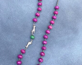May include: A long necklace featuring faceted magenta beads interspersed with small metallic beads. The design includes two silver-toned hand-shaped charms positioned on either side of a single, round, bright green bead, creating a focal point.