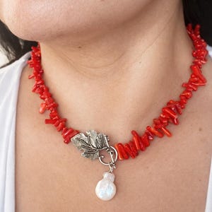 Puede incluir: Un collar de coral rojo vibrante con un cierre plateado en forma de hoja y un colgante de perla blanca. El collar est&aacute; compuesto por peque&ntilde;as piezas de coral irregulares, creando un contraste llamativo.