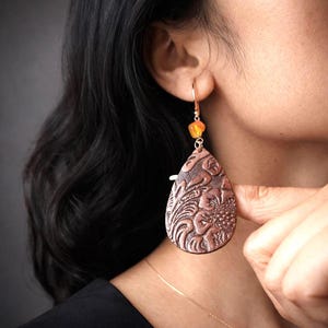 以下が含まれることがあります： 銅色のしずく型のイヤリングで、花のエンボス加工が施されています。イヤリングには、小さなオレンジ色のビーズと金色のフックが付いています。イヤリングは人が着用しています。