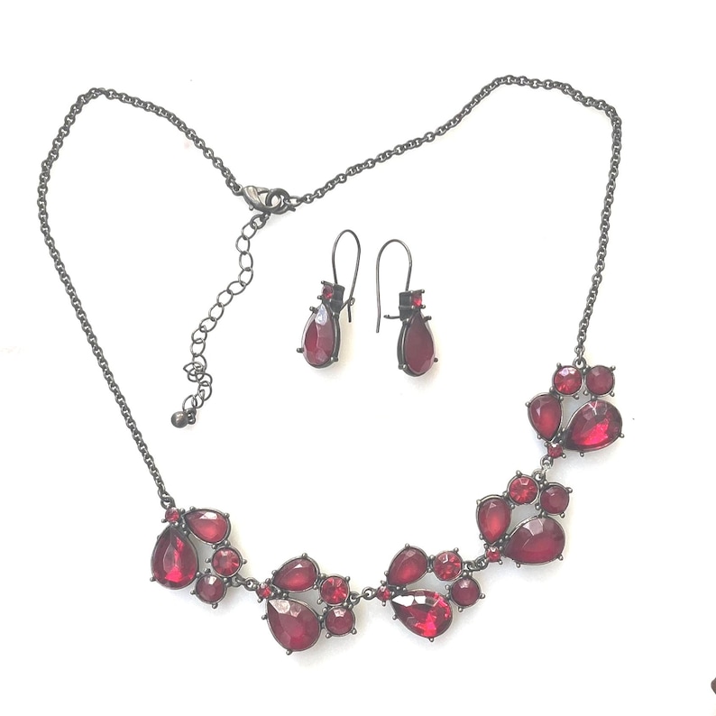 Pu&ograve; includere: Un set di collana e orecchini con catene e montature in metallo grigio scuro. La collana presenta un gruppo di pietre rosse a forma di goccia. Gli orecchini hanno una singola pietra rossa a forma di goccia. Il set di gioielli &egrave; esposto su sfondo bianco.