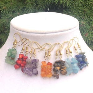Pode incluir: Uma coleção de brincos pendentes com grupos de pedras preciosas brutas e polidas em várias cores, incluindo verde, vermelho, roxo, laranja, castanho, azul claro e azul escuro, suspensos por ganchos de metal dourado.
