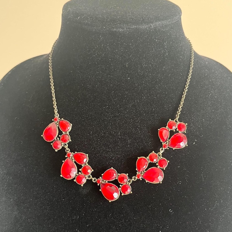 Pu&ograve; includere: Una collana appariscente con gruppi di pietre rosse a forma di goccia. Le pietre sono incastonate in un metallo dorato, creando un design audace. La collana &egrave; esposta su un manichino nero.