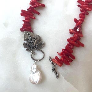 Puede incluir: Un collar de coral rojo con un colgante de hoja plateada y una perla grande e irregular. El collar est&aacute; compuesto por peque&ntilde;as cuentas de coral cil&iacute;ndricas. El colgante es un dise&ntilde;o de hoja plateada detallada.