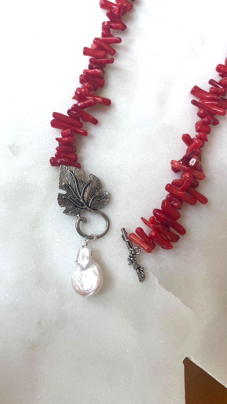 Puede incluir: Un collar de coral rojo con un colgante de hoja plateada y una perla grande e irregular. El collar est&aacute; compuesto por peque&ntilde;as cuentas de coral cil&iacute;ndricas. El colgante es un dise&ntilde;o de hoja plateada detallada.