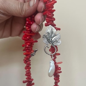 Puede incluir: Un collar de coral rojo con un colgante de hoja plateada y una perla blanca. El collar est&aacute; hecho de peque&ntilde;os trozos de coral irregulares. El colgante de hoja tiene detalles intrincados.