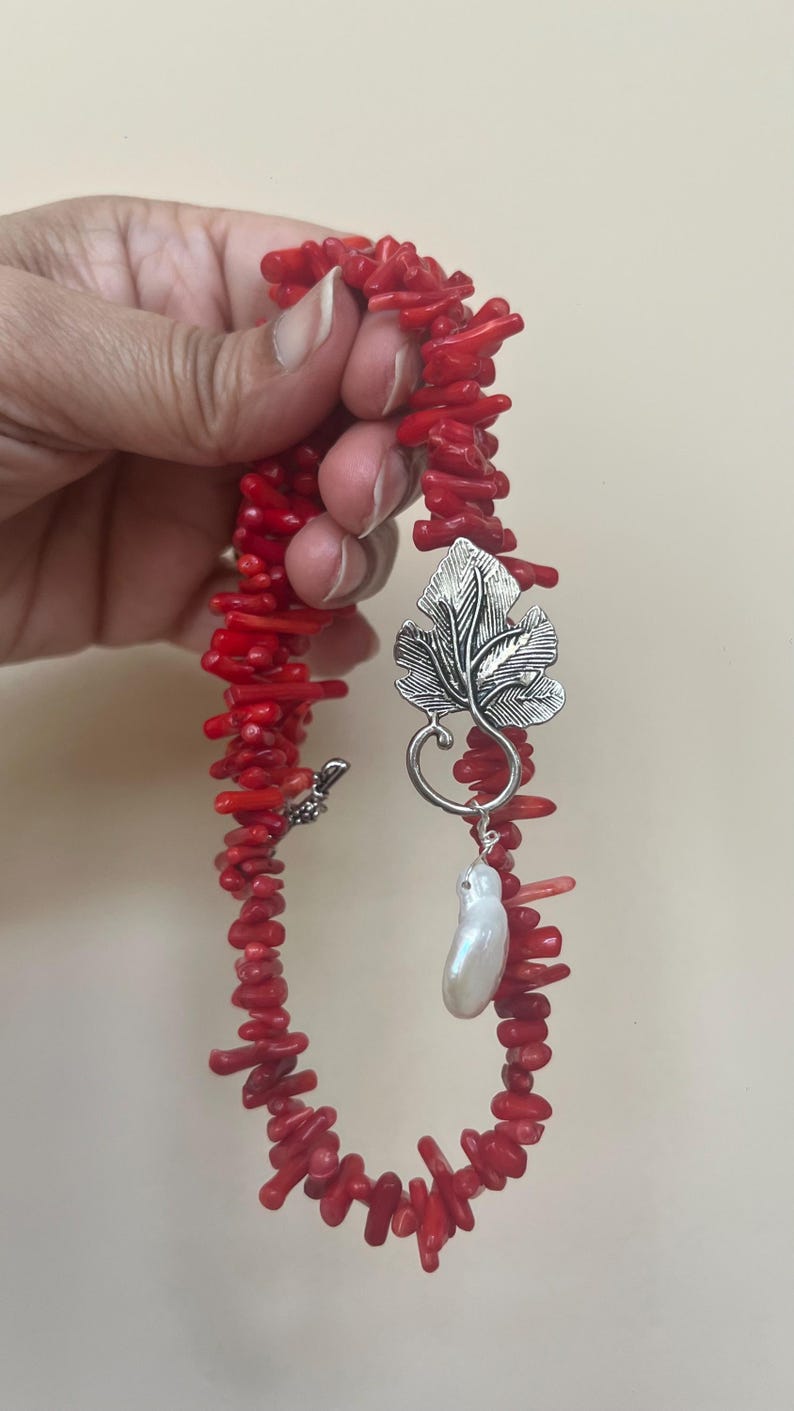 Puede incluir: Un collar de coral rojo con un colgante de hoja plateada y una perla blanca. El collar est&aacute; hecho de peque&ntilde;os trozos de coral irregulares. El colgante de hoja tiene detalles intrincados.
