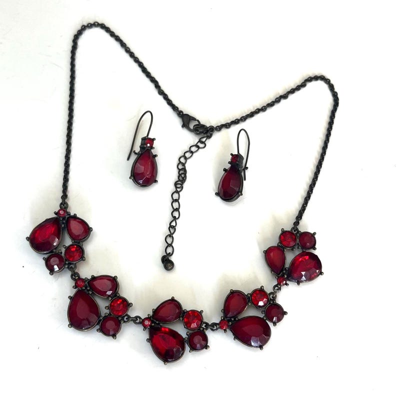 Pu&ograve; includere: Un set di collana e orecchini in metallo canna di fucile scuro. La collana presenta gruppi di pietre preziose rosse a forma di goccia e rotonde. Gli orecchini abbinati hanno una singola pietra preziosa rossa a forma di goccia. Il set di gioielli &egrave; esposto su sfondo bianco.