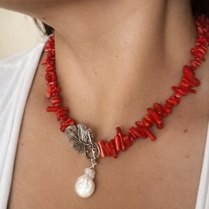 Puede incluir: Un collar de coral rojo con un cierre de hoja plateada y un colgante de perla blanca. El collar est&aacute; compuesto por peque&ntilde;os trozos irregulares de coral. El cierre plateado tiene un dise&ntilde;o de hoja detallado. La perla tiene forma de l&aacute;grima.