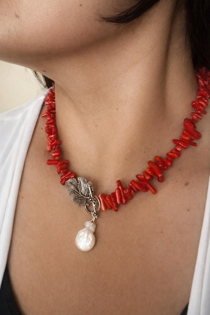 Puede incluir: Un collar de coral rojo con un cierre de hoja plateada y un colgante de perla blanca. El collar est&aacute; compuesto por peque&ntilde;os trozos irregulares de coral. El cierre plateado tiene un dise&ntilde;o de hoja detallado. La perla tiene forma de l&aacute;grima.