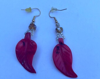 May include: A pair of dangle earrings featuring bright red leaf-shaped pendants suspended from gold-toned metal hooks. Each earring includes a small faceted brown bead and a cluster of tiny clear seed beads connecting the hook to the leaf.