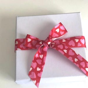 Pu&ograve; includere: Una scatola regalo quadrata bianca legata con un nastro rosso a fantasia di cuori rosa e bianchi. Il nastro &egrave; annodato a fiocco sulla parte superiore della scatola, creando una presentazione festosa e romantica.