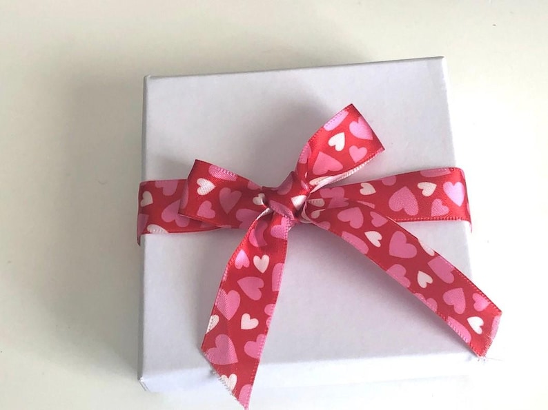 Pu&ograve; includere: Una scatola regalo quadrata bianca legata con un nastro rosso a fantasia di cuori rosa e bianchi. Il nastro &egrave; annodato a fiocco sulla parte superiore della scatola, creando una presentazione festosa e romantica.