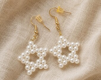 May include: A pair of dangle earrings featuring star-shaped charms constructed from small, round, white faux pearls. The earrings are attached to gold-toned metal fishhook wires and include clear plastic earring backs for secure placement.