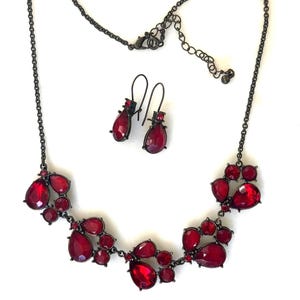 Pu&ograve; includere: Un set di gioielli composto da collana e orecchini. La collana e gli orecchini hanno una finitura in metallo scuro e sono adornati con pietre preziose rosse a forma di goccia. La collana ha un design a grappolo, e gli orecchini sono semplici pendenti.