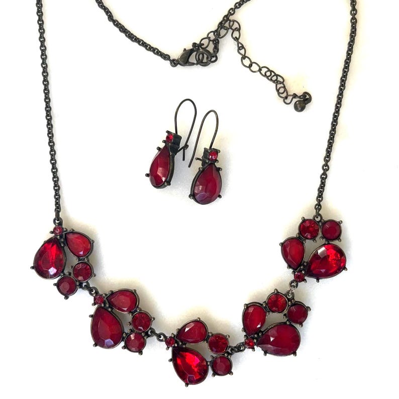 Pu&ograve; includere: Un set di gioielli composto da collana e orecchini. La collana e gli orecchini hanno una finitura in metallo scuro e sono adornati con pietre preziose rosse a forma di goccia. La collana ha un design a grappolo, e gli orecchini sono semplici pendenti.
