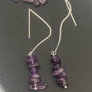 Puede incluir: Un par de pendientes de hilo en tono plateado con racimos colgantes de piedras preciosas de amatista morada, pulidas e irregulares. Cada pendiente consta de una fina cadena metálica con un poste recto y una disposición vertical de piedras.