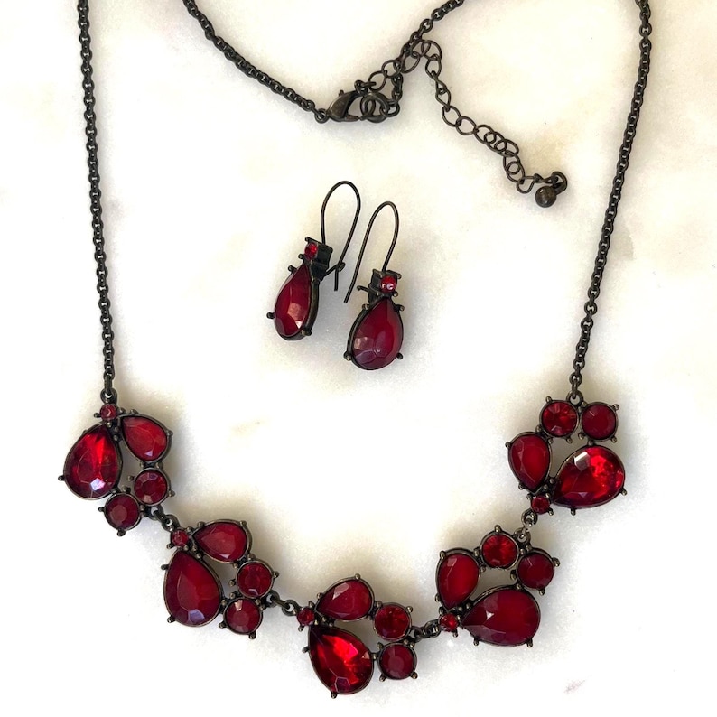 Pu&ograve; includere: Un set di collana e orecchini in metallo canna di fucile scuro con grappoli di pietre preziose rosse a forma di goccia e rotonde. La collana ha una catena regolabile. Gli orecchini hanno un design a goccia abbinato. Il set di gioielli &egrave; esposto su una superficie bianca.