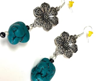 May include: A pair of dangle earrings featuring a silver-toned metal flower with intricate filigree cutouts, suspended from a hook. Below the flower, a single irregular, smooth turquoise-colored stone is accented by small black and white beads.